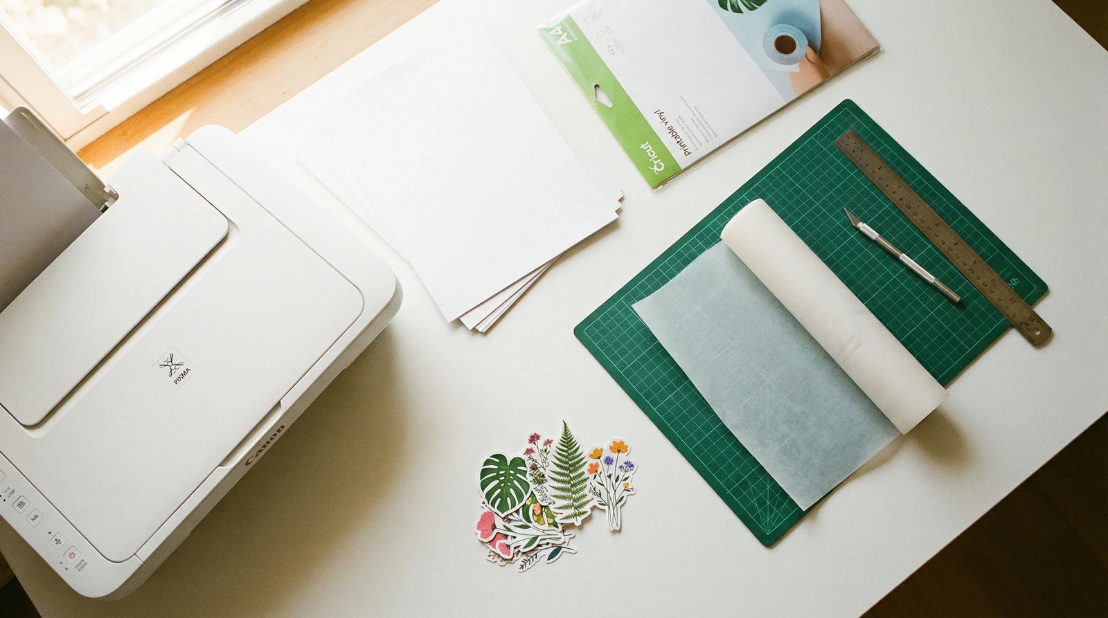 Sticker business startup supplies including printer, vinyl sheets, cutting mat, release paper, and finished die-cut plant stickers on a white desk